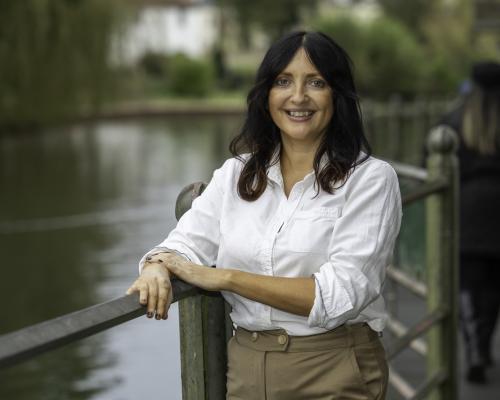 Donna - A female with should length dark hair, smiling against the railing. They are wearing a white shirt and beige trousers. 