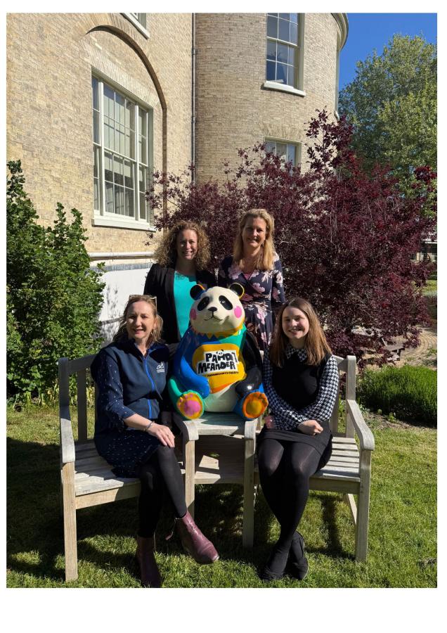 A group of four women, two standing behind and two sitting on a bench with a panda sculpture between them. The setting is the grounds of a manor house.
