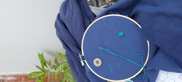 An embroidery hoop holding a blue hoody top, which has been used to practice sewing on a button, mending a hole using satin stitch and basic mending techniques, that Natalie completed during our workshop.