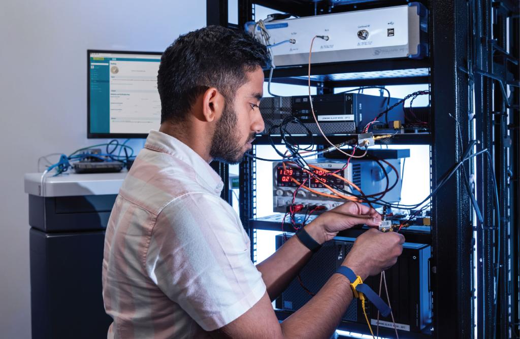 An engineer testing connected medical device hardware in a laboratory. He is surrounded by wires and screens and holds some cables to the right hand side of the image. 