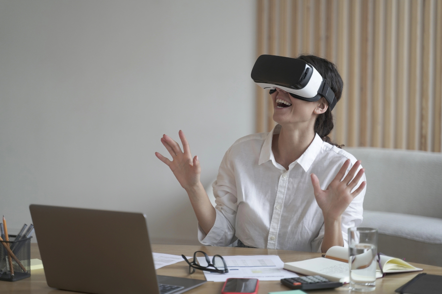 Business lady sits at work desk wearing a VR headset for laptop, raising her hands up as if trying to touch objects in 3D reality
