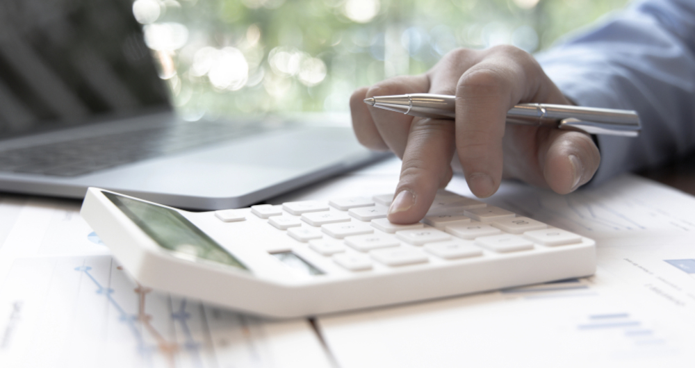 Accountant's hand on a calculator ,with laptop alongside, calculating income expenditure and investment data