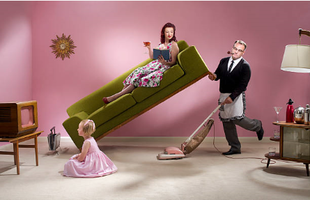 A man in a apron, lifting a sofa with to clean. Mother sitting and reading a book while a little girl watches TV 