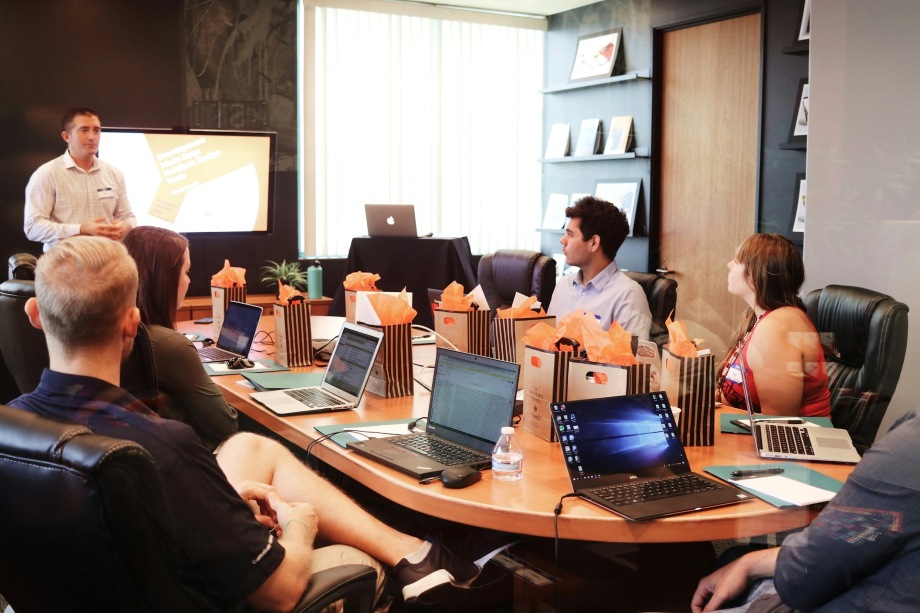 A group of people sitting around a large table in an office, with a trainer standing at the end of the table in front of a large wall-mounted screen