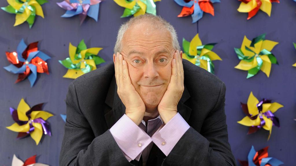 Gyles Brandreth resting his face in his hands. Purple background with windmills on it. 