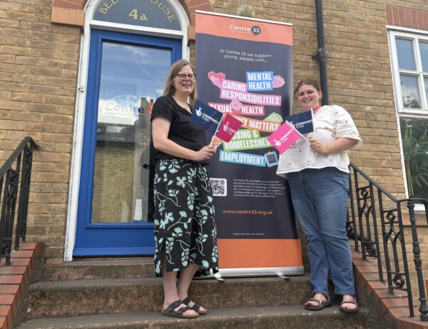 Harriet Millward from Centre 33 and Tia Halliday from the National Lottery Community Fund, smiling on the steps outside Centre 33's new building and holding National Lottery Community Fund promotional materials. Behind them is a Centre 33 banner highlighting support services for young people, including mental health, caring responsibilities, sexual health, and employment.
