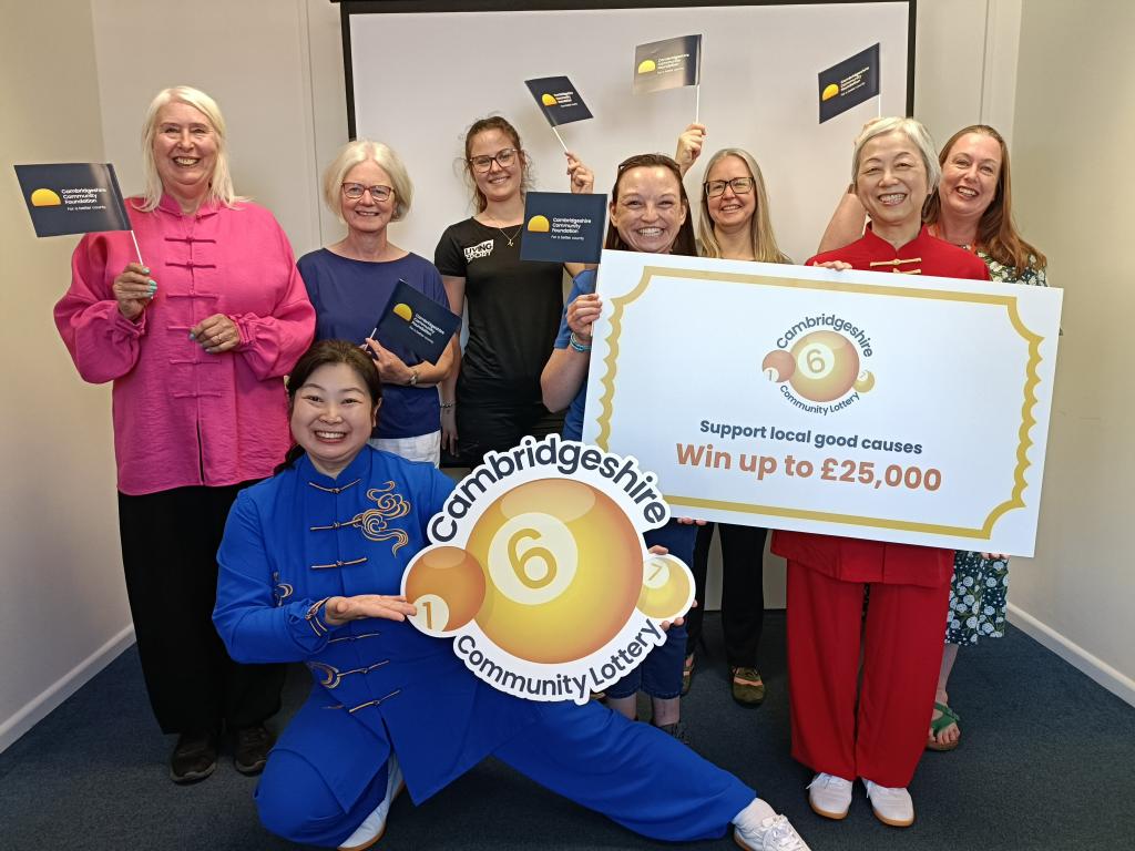 Charity representatives standing together holding signs with the Cambridgeshire Community Lottery logo and a branded lottery ticket.
