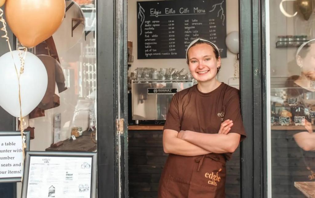 Woman leaning on the door frame of her cafe, smiling at the camera.