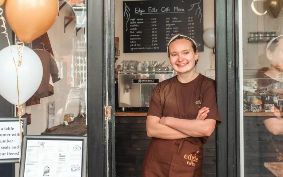 Woman leaning against the door frame of her cafe, smiling at the camera.