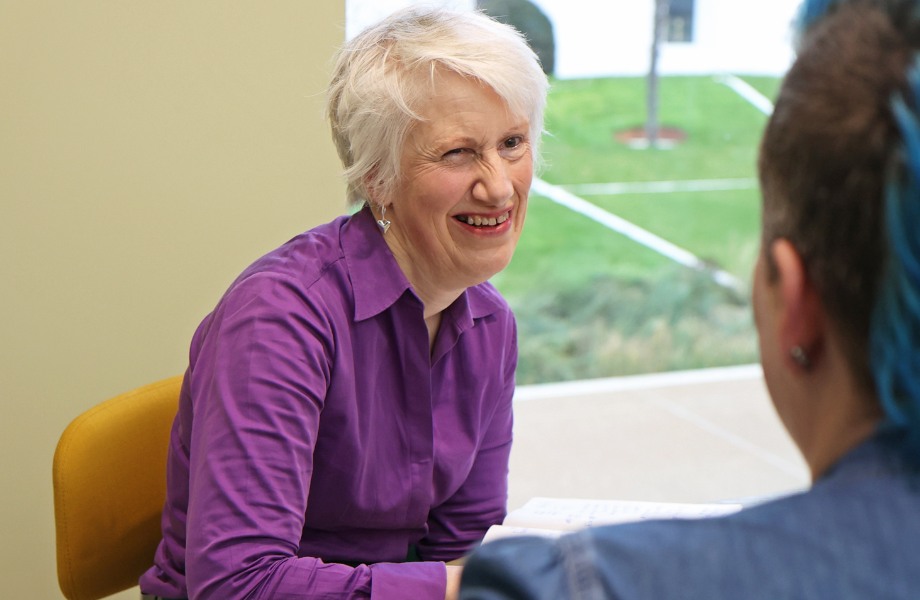 Sarah Stones is sitting at a table wearing a purple shirt and smiling at a client who is just visible to the right-hand side of the shot.