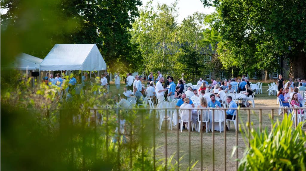 guests network and mingle, some standing and some seated around white tables, outdoors on the jesus college orchard lawn on a warm summer evening 