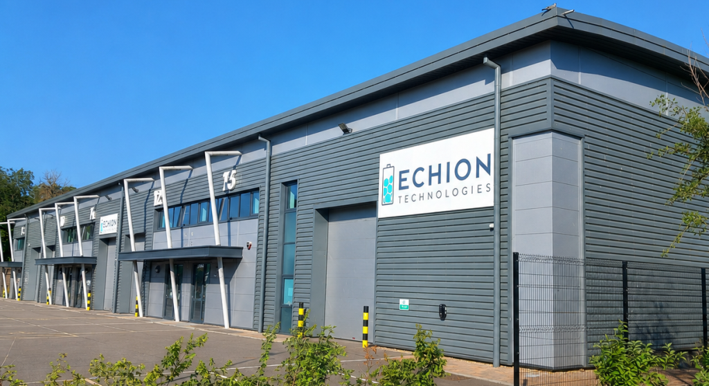 Exterior photograph of an Echion Technologies facility on a bright, clear day. The modern grey industrial building has large roller doors and several entrance bays, with the Echion Technologies logo displayed prominently on the front. The building is set within a paved business park, with trees and greenery around the site.