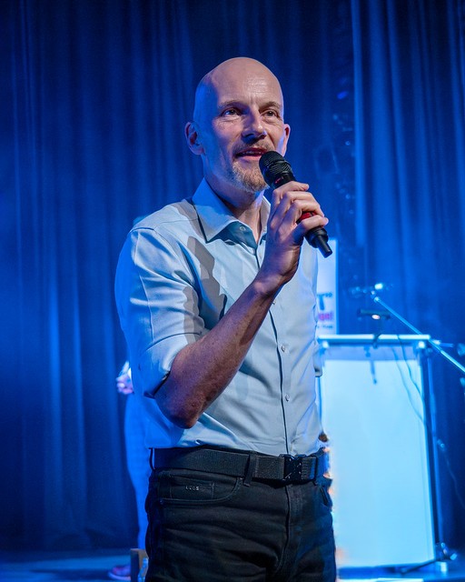 A bald man standing and speaking while holding a microphone.