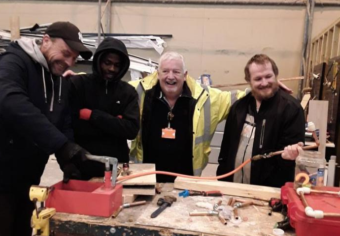Image of plumbing tutor and 3 participants, posing for an image, smiling and laughing, around a work table in a workshop