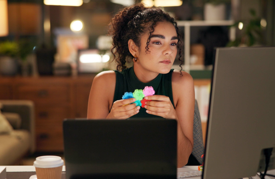 Remote worker - a female with a black tank top sits at their desk with a laptop open. They are staring at a computer screen and are playing with a fidget toy.