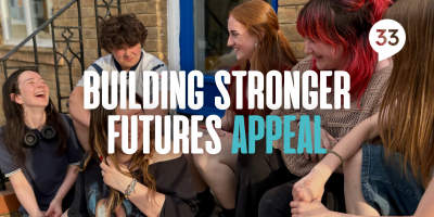 A group of young people sitting on the steps of the new Centre 33 building, laughing and smiling, with the text "Building Stronger Futures Appeal" over the top