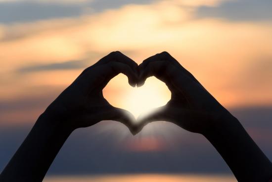 Two hands togehter forming a love heart shape with a sunset background 