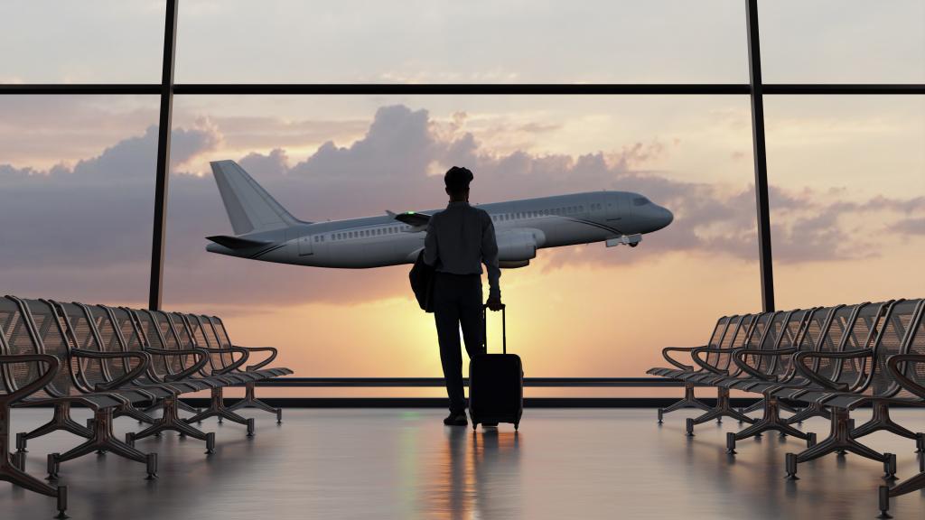 A man stands in an airport, looking out of a window as a plane takes off in the distance 