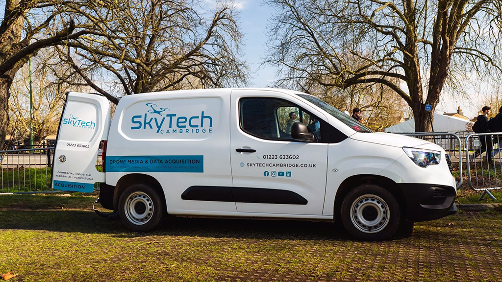 White SkyTech van parked on grass, back door open. Park setting, trees in background.