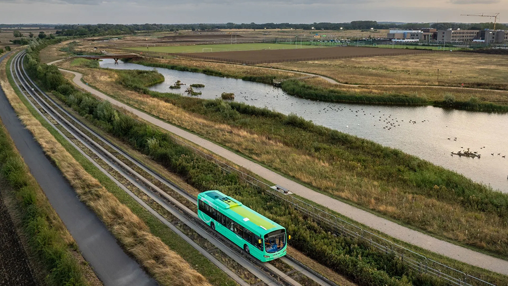 Green bus travels along the Cambridge guided busway tracks through a rural landscape next to Northstowe and its new lakes