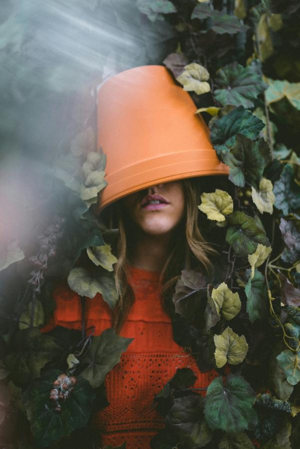 A young woman with a flowerpot on her head that obscures her eyes. She is immersed in leaves and vines.