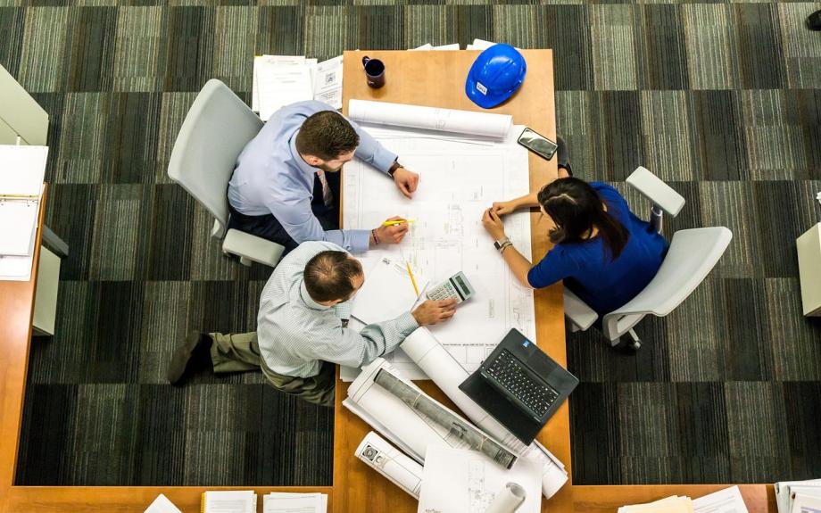 Two men and a woman sitting at a desk which has blueprints and a hard hat on it.