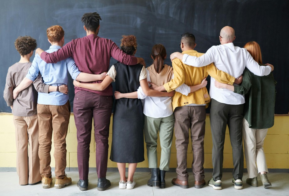 A diverse group of people standing with their backs to the camera and arms around each others' waists.
