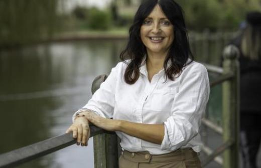 Donna - A female with should length dark hair, smiling against the railing. They are wearing a white shirt and beige trousers. 