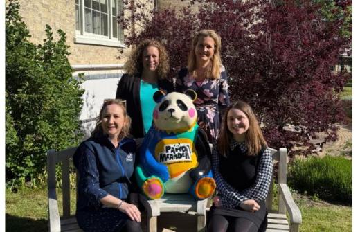 A group of four women, two standing behind and two sitting on a bench with a panda sculpture between them. The setting is the grounds of a manor house.