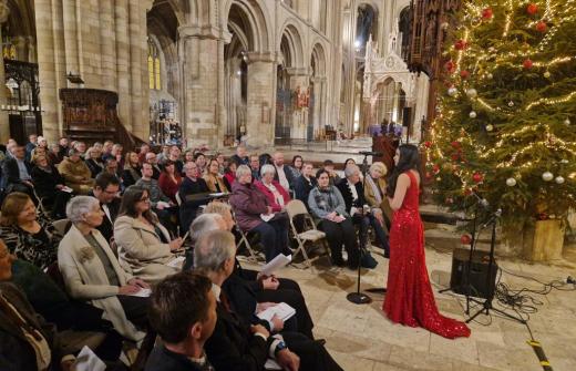 Singer, Gabriella Pineda-Rodrigues, in a red sequined dress performing in front of guests inside Peterborough Cathedral. A decorated Christmas tree is next to the singer.