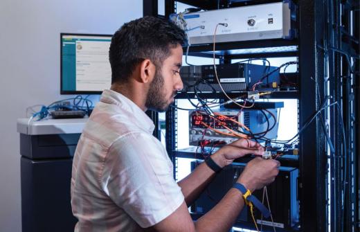 An engineer testing connected medical device hardware in a laboratory. He is surrounded by wires and screens and holds some cables to the right hand side of the image. 