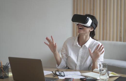 Business lady sits at work desk wearing a VR headset for laptop, raising her hands up as if trying to touch objects in 3D reality