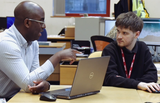 A man with glasses and a striped shirt points toward a laptop screen while speaking to a young man wearing a "VISITOR" lanyard in an office setting.