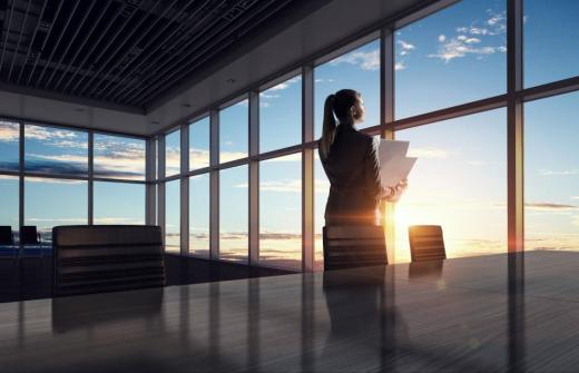 Women in corporate setting looking out office building at sunrise with paperwork in hand.