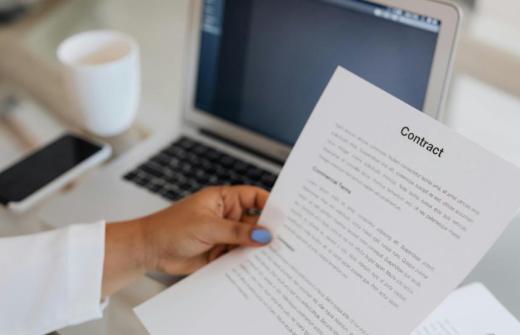 A hand holding a contract over a laptop on a desk.