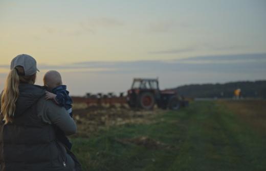 Mother holds baby while overlooking a tractor in field at sunset 