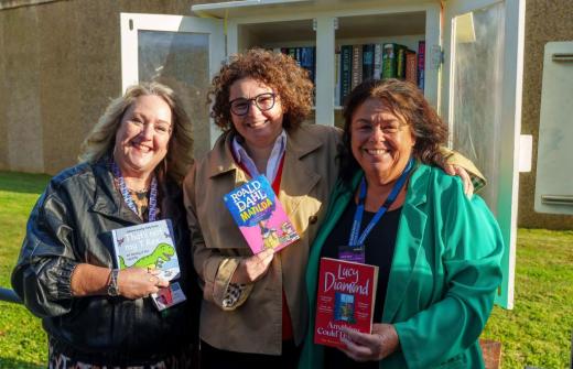 Three women stood in front of Little Free Library holding books