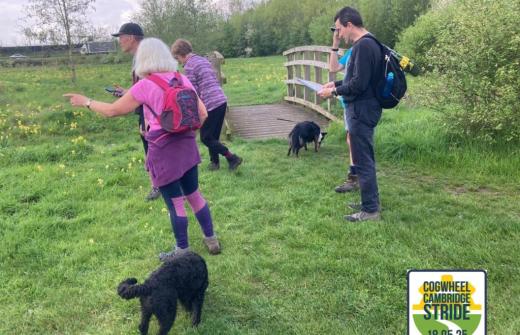 The image shows a small group of people with two dogs, walking across a field as part of the Cogwheel Cambridge Stride in 2023.
