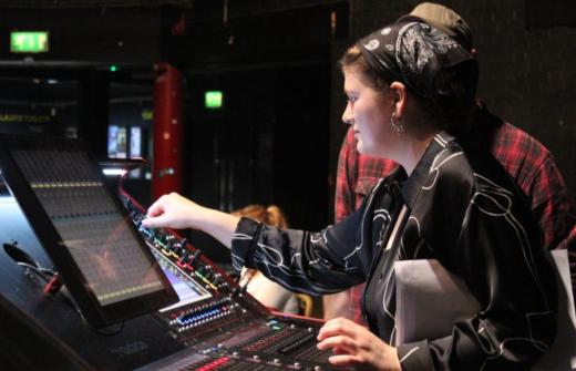 A young woman wearing a black shirt is at a mixing desk, adjusting the controls.