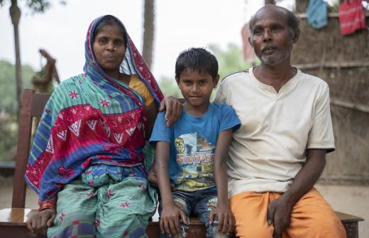 Aanand, wearing a blue t-shirt sits between his parents outside. His mother is resting her hand on his shoulder.