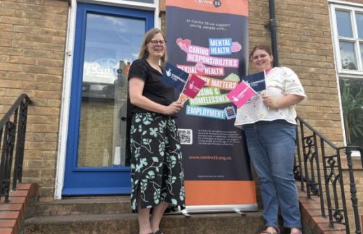Harriet Millward from Centre 33 and Tia Halliday from the National Lottery Community Fund, smiling on the steps outside Centre 33's new building and holding National Lottery Community Fund promotional materials. Behind them is a Centre 33 banner highlighting support services for young people, including mental health, caring responsibilities, sexual health, and employment.