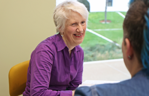 Sarah Stones is sitting at a table wearing a purple shirt and smiling at a client who is just visible to the right-hand side of the shot.