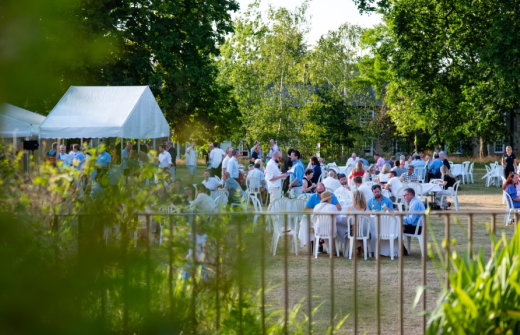 guests network and mingle, some standing and some seated around white tables, outdoors on the jesus college orchard lawn on a warm summer evening 