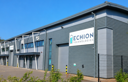 Exterior photograph of an Echion Technologies facility on a bright, clear day. The modern grey industrial building has large roller doors and several entrance bays, with the Echion Technologies logo displayed prominently on the front. The building is set within a paved business park, with trees and greenery around the site.