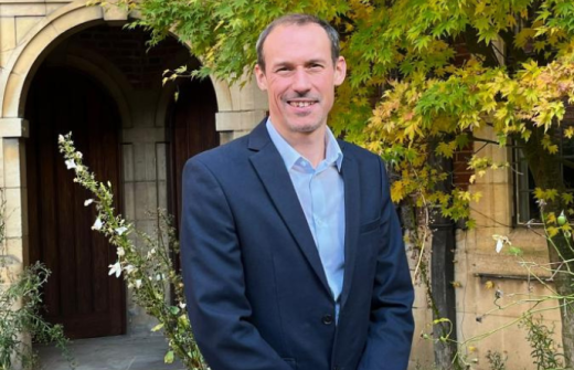 White man in a dark blue suit smiling at the camera outside in front of an old Cambridge College with greenery in the background. Andrew Bell the new Head of Meet Cambridge