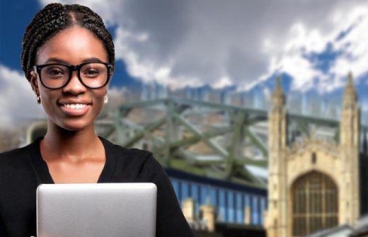 A smiling lady standing with a laptop. The bridge of sighs, Kings college and the Fitzwilliam Museum are in the background