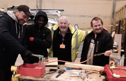 Image of plumbing tutor and 3 participants, posing for an image, smiling and laughing, around a work table in a workshop