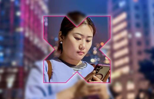 A woman looks intently at her smartphone while standing in a city at night. The scene is overlaid with a bold pink-outlined “X” shape, sharply focused on the woman while the surrounding city lights and buildings are blurred — suggesting focus on digital connection or social media in an urban context.