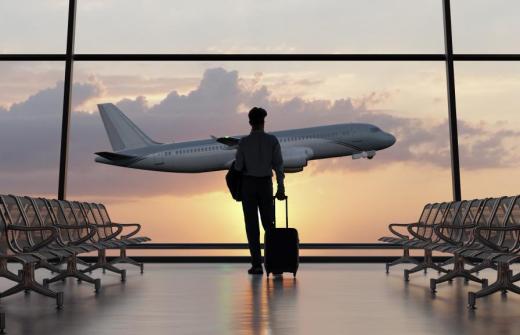 A man stands in an airport, looking out of a window as a plane takes off in the distance 