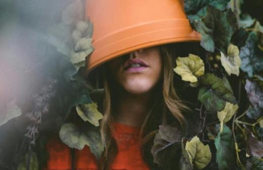A young woman with a flowerpot on her head that obscures her eyes. She is immersed in leaves and vines.